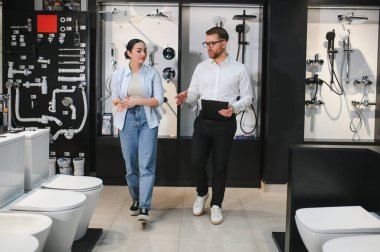 Satıcı kadın müşterilere farklı banyo armatürleri gösteriyor. Tesisat ve seramik seramik dükkanlarında dolaşıyorlar.