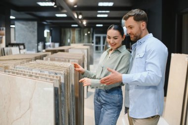 Çift modern bir mağazada çeşitli seramik fayanslar inceleyerek, ev yenileme veya iyileştirme projeleri seçeneklerini tartışıyor