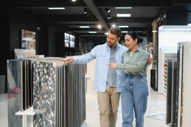 Çift seramik fayans örneklerini gösteriyor, modern bir tesisat dükkanında banyo yenileme malzemeleri hakkında tartışıyor ve seçiyorlar.