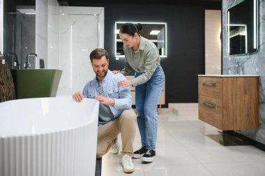 Tesisat ve seramik fayans dükkanında beyaz küveti seçen mutlu çift, karar vermede kocasını destekleyen eş, yeni banyo mobilyası alan aile.