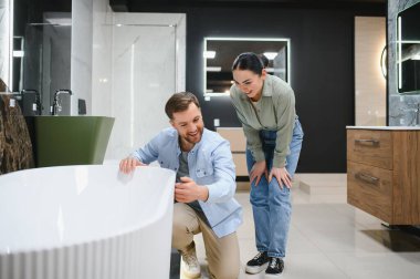 Çift, seramik bir fayans ve su tesisatı dükkanında modern bir küveti inceliyor. Ev yenileme ve iç dekorasyona ilgi gösteriyor.