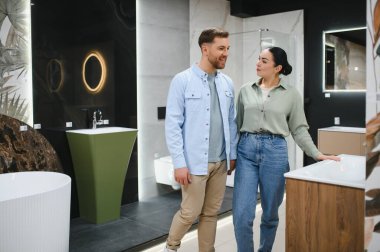 Genç çift, bir su tesisatçısı dükkanında banyo eşyasına el ele tutuşup tartışıyor ve gülümsüyor.