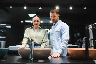 Fayans ve tesisat dükkanında seramik lavaboları inceleyen çift, ev yenileme fikirleri ve banyo yenileme seçeneklerini tartışıyor.