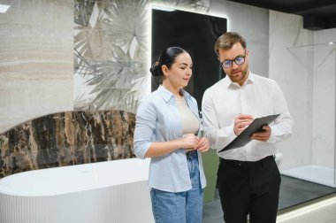 Satış elemanı, müşteriye bir su tesisatı dükkanında banyo malzemeleri seçmesinde yardımcı olurken not alıyor.