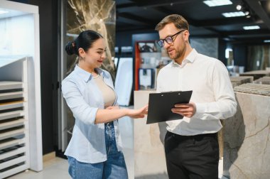 Modern bir seramik ve tesisat mağazasında pano tutan kadın müşterilere farklı kaplama seçenekleri sunan profesyonel bir satıcı.