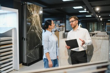 Satıcı elinde bir pano tutarken modern bir inşaat malzemeleri mağazasında bir kadına seramik fayanslar gösteriyor ve çeşitli seçenekleri tartışıyor.