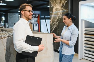Gülümseyen satıcı elinde bir pano var. Tesisat ve fayans dükkanındaki mutlu bayan müşteriye seramik fayanslar gösteriyor.