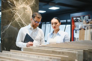 Satıcının elinde pano var. Tesisat ve kiremitçide müşteriye seramik fayans örnekleri gösteriyor.