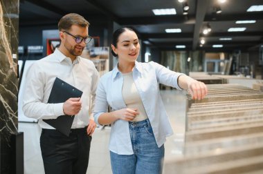 Hırdavatçıda seramik fayans seçen kadın müşteri, elinde pano tutan satıcı ona yardım ediyor.