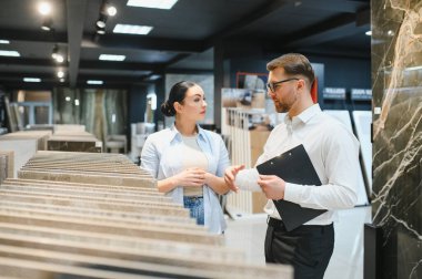 Satıcının elinde bir pano seramik ve sıhhi tesisat dükkanındaki müşteriye seramik fayanslar gösteriyor.