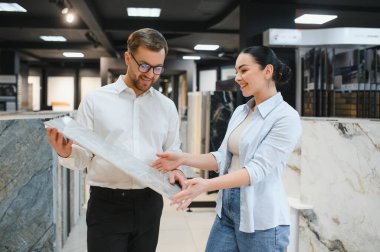 Gülümseyen tezgahtar hırdavatçıdaki kadın müşteriye seramik fayanslar gösteriyor. Dairesini yenilemek için malzeme seçiyor.