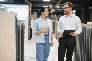 Satış elemanı elinde seramik seramik seramik koleksiyonu, hırdavatçı dükkanının koridorunda yürüyen kadın müşteriye gösteriyor.
