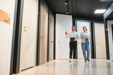 Satıcı hırdavatçıda bir kadına farklı kapı modelleri gösteriyor, hangisini seçeceğini öneriyor.