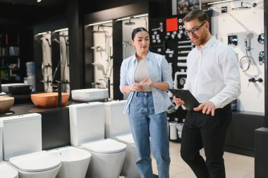 Satıcı elinde farklı tuvalet modellerini modern banyo eşyaları mağazasındaki müşterilere gösteriyor.