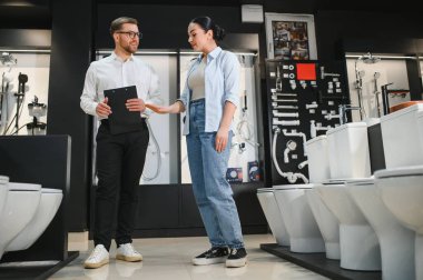 Satıcının elinde pano var. Tuvalet kâselerini kadın müşterilere gösteriyor. Tesisat ve banyo mobilyası mağazasında.