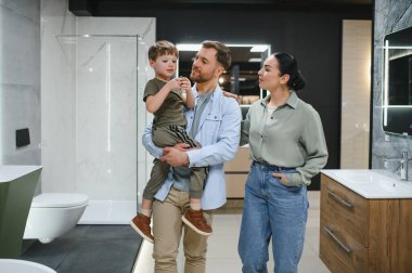 Aile, tesisat dükkanında yeni banyo eşyaları seçiyor, baba oğlunu kollarına alıyor, anne ürünlere bakıyor.