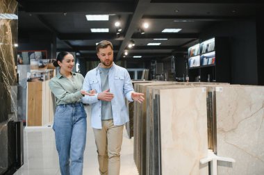 Genç bir çift, banyolarını yenilemek için seramik fayanslar seçerek bir hırdavatçıda yürüyor, farklı örnekleri tartışıyor ve işaret ediyor.