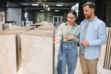 Çift modern bir mağazada seramik fayansları inceliyor, ev yenileme seçeneklerini tartışıyor. Dekor, çeşitli sergileri olan iyi aydınlatılmış bir sergi salonu.