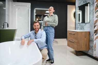 Gülümseyen çift seramik küveti seçiyor. Su tesisatı ve banyo malzemeleri mağazasında. Kadın gülüyor, erkek küvetin yanında çömelmiş.