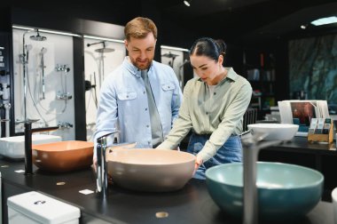 Çift modern bir sergi salonunda seramik lavaboları ve tesisat tesisatlarını inceliyor, ev geliştirme ve iç tasarım seçeneklerini sergiliyor.