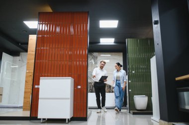 Satış elemanı, elinde bir pano olan ve ürünlere işaret eden kadın müşteriye farklı banyo fayansları ve eşyalar gösteriyor.