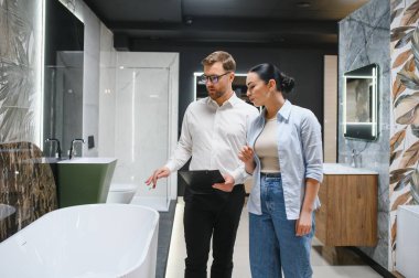 Profesyonel tezgahtar, tesisat dükkanındaki müşteriye farklı banyo armatürlerini gösteriyor, özellikleri ve faydaları tartışıyor.