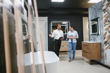 Çağdaş bir mağaza ortamında seramik fayans ve tesisat seçeneklerini araştıran kadın alıcı ve erkek satıcı, modern banyo tasarımlarını ön plana çıkarıyor.