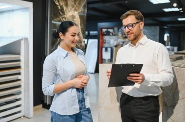 Kadın müşteriye seramik örnekler gösteren gülümseyen bir satıcı elinde bir pano tutuyor ve satın alma seçeneklerini tartışıyor.