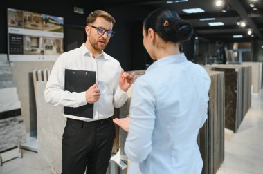 Parlak bir tesisat ve fayans dükkanında kadın müşteriye çeşitli seramik fayans örnekleri sunan satıcı, satın alma kararını kolaylaştırıyor.