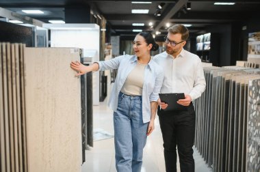 Müşteri seramik ve su tesisatında elinde pano tutan bir satıcıyla konuşurken seramik bir fayans örneğini işaret ediyor.