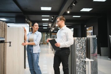 Seramik ve sıhhi tesisat dükkanında elinde büyük bir fayans örneği tutan bir kadına farklı seramik fayans örnekleri gösteren bir tezgahtar.