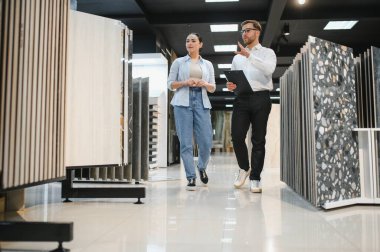 Modern bir tesisat ve fayans dükkanının vitrininde yürüyen bir kadına farklı seramik fayanslar gösteren profesyonel bir satıcı.