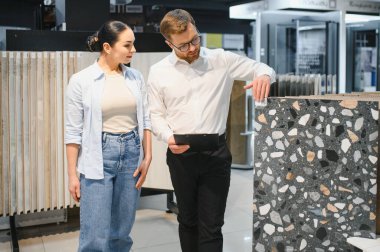 Satıcı, bir ev geliştirme mağazasında alışveriş yaparken çeşitli seramik fayanslar seçen bir kadına yardım ediyor ve yenileme projesi için seçenekleri araştırıyor.