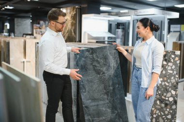 Modern ev geliştirme mağazasındaki gülümseyen kadın müşterilere koyu mermer seramik örneği sunar.