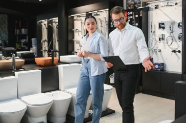 Profesyonel satıcı, kadın müşteriye banyo malzemeleri sunuyor, seçenekleri tartışıyor ve modern bir tesisat mağazasında uzman tavsiyesi veriyor.