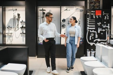 Satış elemanı, tesisat dükkanındaki kadın müşterilere çeşitli banyo demirbaşlarını gösteriyor, yenileme ve iyileştirme seçeneklerini tartışıyor.