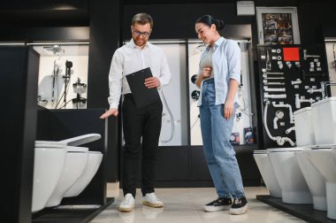 Tesisatçı dükkanında bir kadına farklı klozetler gösteren gülümseyen bir satıcı. İhtiyaçları için doğru olanı seçmesine yardım ediyor.