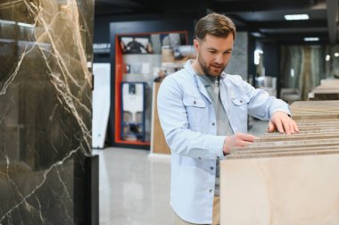 Hırdavatçıdan seramik fayanslar seçen, numunelere dokunan ve yenileme projesi için mükemmel malzemeyi arayan bir adam.