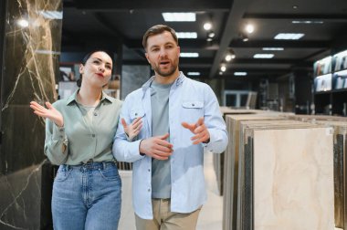 Çift, çeşitli fayans örnekleri ve modern eşyalar ile çevrili seramik bir fayans ve tesisat mağazasını keşfederken seçenekleri tartışıyor.