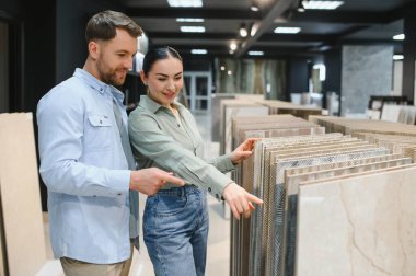 Gülümseyen çift seramik fayans örneklerine işaret ediyor, modern bir hırdavatçıda banyo veya mutfak yenileme malzemeleri seçiyor