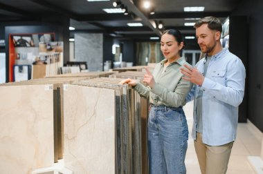 Modern bir tesisat ve fayans dükkanında seramik fayansları inceleyen çift, çeşitli fayanslarla çevrili ev yenileme için tasarım seçeneklerini tartışıyor.