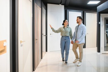 Genç bir çift el ele tutuşup hırdavatçıda yeni iç kapılar seçiyor. Tahta kapıyı gösteren bir kadın.