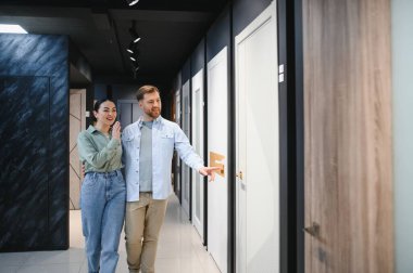 Mutlu çift bir hırdavatçıda yeni iç kapılar seçiyor, beyaz bir kapıya yürüyor ve işaret ediyor, evi yenilemek için alışveriş yapıyor.