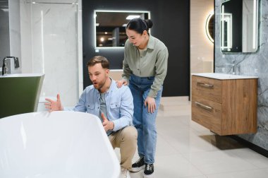 Genç çift banyo mobilyası dükkanında seramik küveti seçiyor, yenileme projesini tartışıyor ve planlıyorlar.