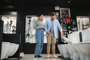 Genç çift, banyo yenileme projeleri için hijyenik malzemelerini incelemek üzere bir su tesisatı dükkanında seramik klozet seçiyor.