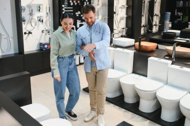 Genç çift gülümseyip tartışırken tesisat mağazasında yeni bir klozet seçiyorlar ve ev geliştirme sürecinin tadını çıkarıyorlar.