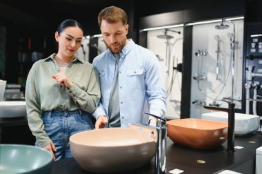 Çift modern banyo tasarım seçeneklerinin sergilendiği çağdaş bir fayans ve tesisat mağazasında şık seramik lavaboları ve muslukları inceliyor.