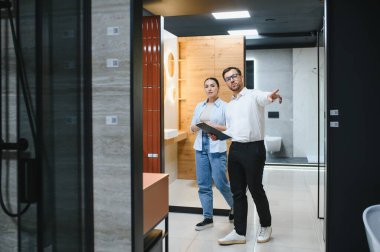 Satıcı, kadın müşteriye seramik fayanslar ve sergi salonundaki tesisatı seçmesinde yardımcı olurken banyo demirbaşlarını işaret ediyor.