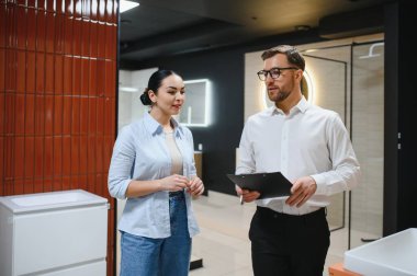Profesyonel satıcı elinde bir pano tutuyor. Tesisatçı dükkanındaki kadın müşterilere farklı banyo malzemelerini gösteriyor.