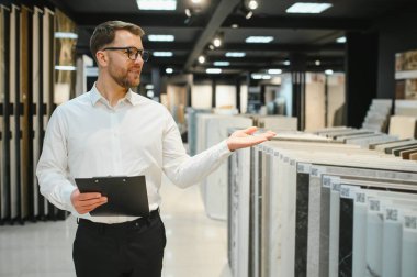 Modern bir hırdavatçıda müşteriye çeşitli seramik fayans örnekleri sunan satıcı, ev geliştirme seçenekleri vurguluyor